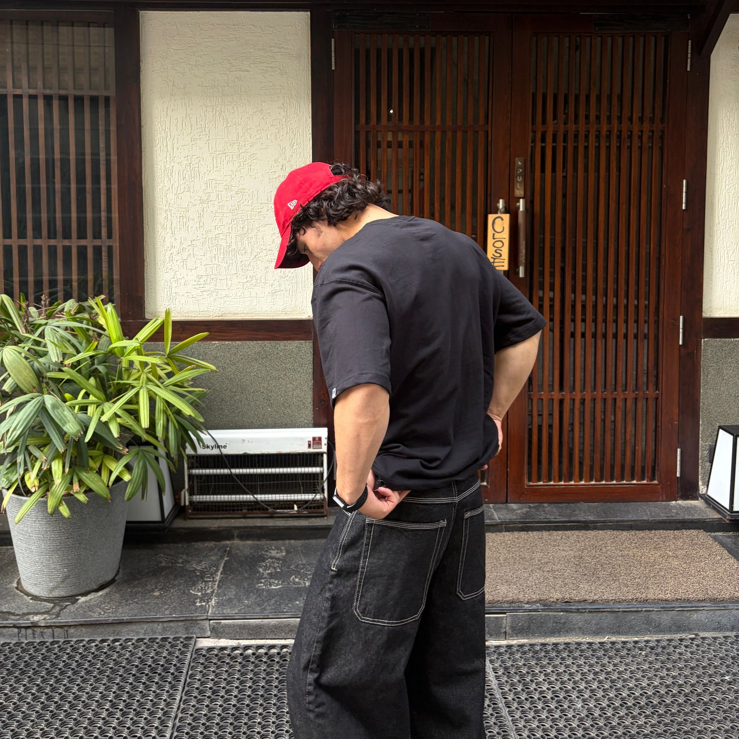 Person wearing a red cap and black shirt standing outside a building with a plant nearby.