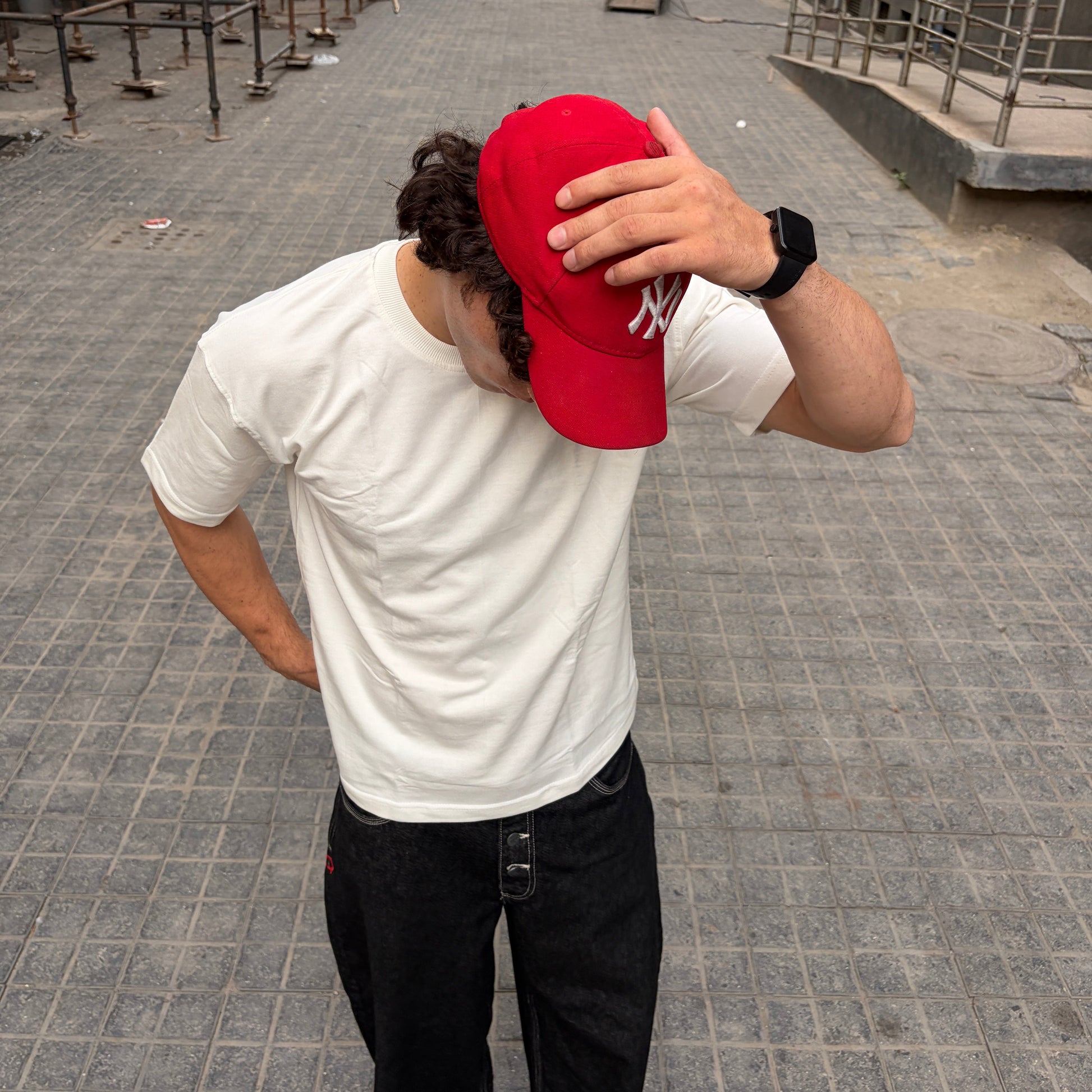 Person wearing an off-white t-shirt and black pants, holding a red cap over their head on a city street.