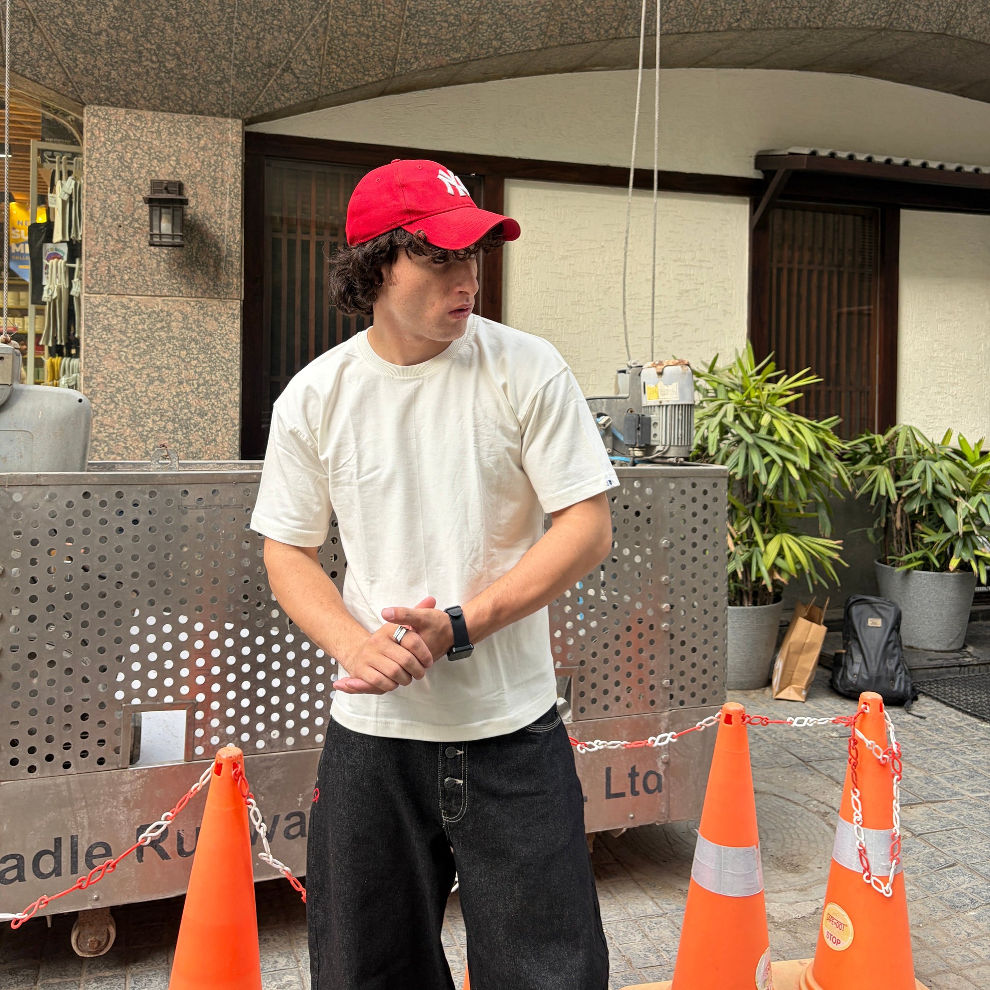 Person wearing a red cap and off-white shirt standing on a sidewalk with orange traffic cones nearby.