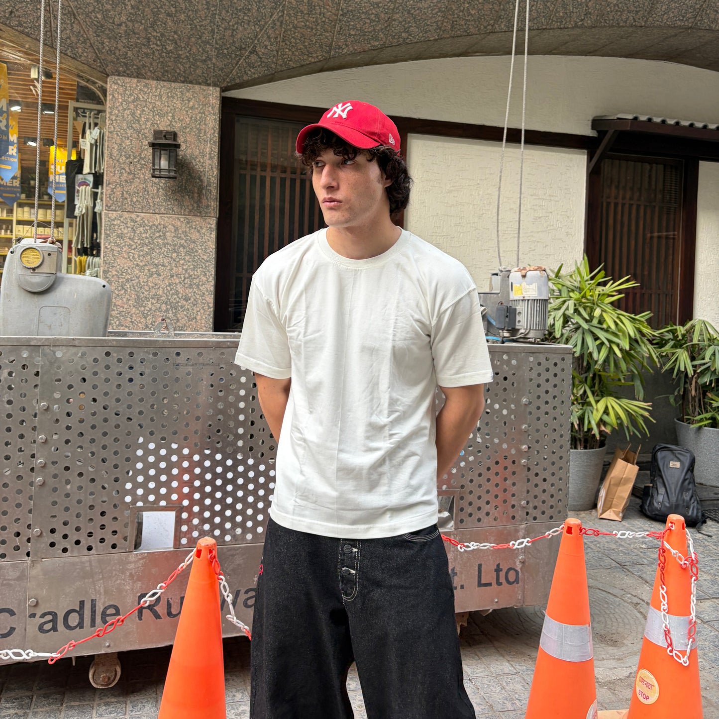 Person wearing a red cap and off-white t-shirt standing between orange traffic cones outdoors.