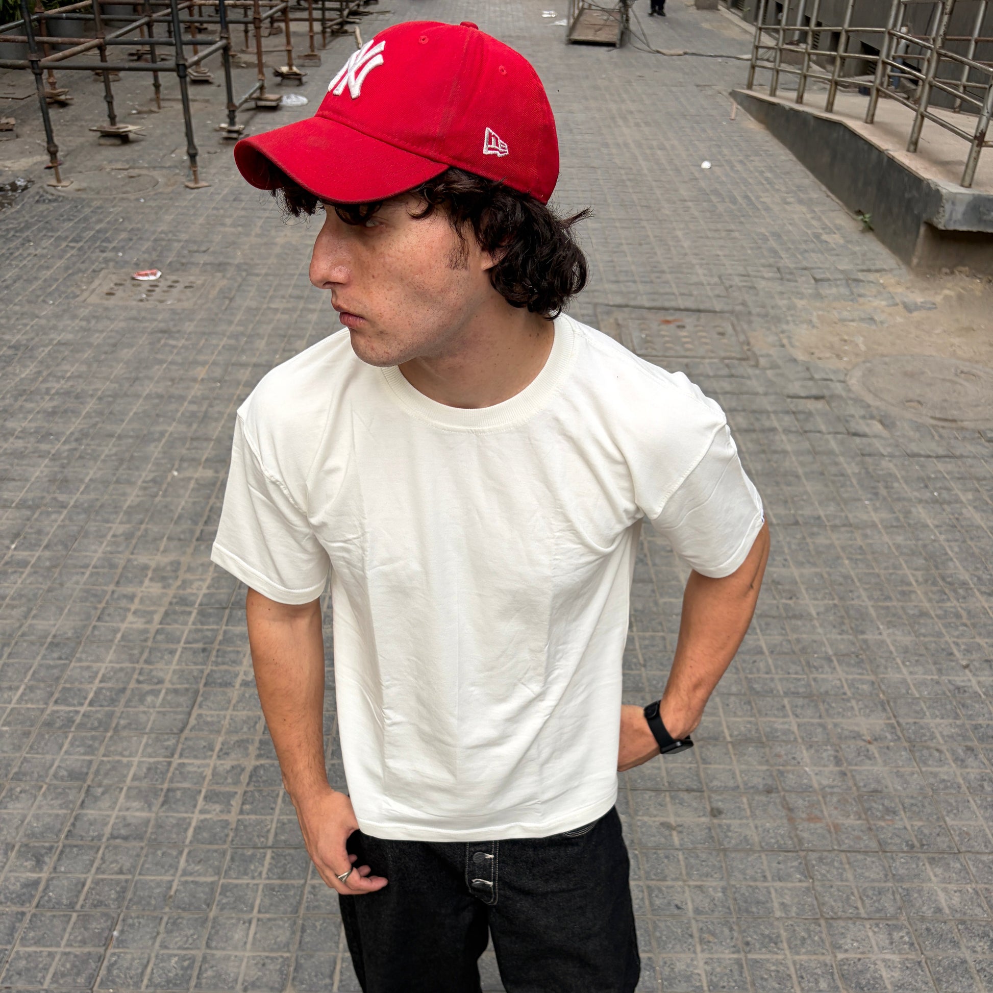 Person wearing a off-white t-shirt, black pants, and a red cap on a gray pavement.