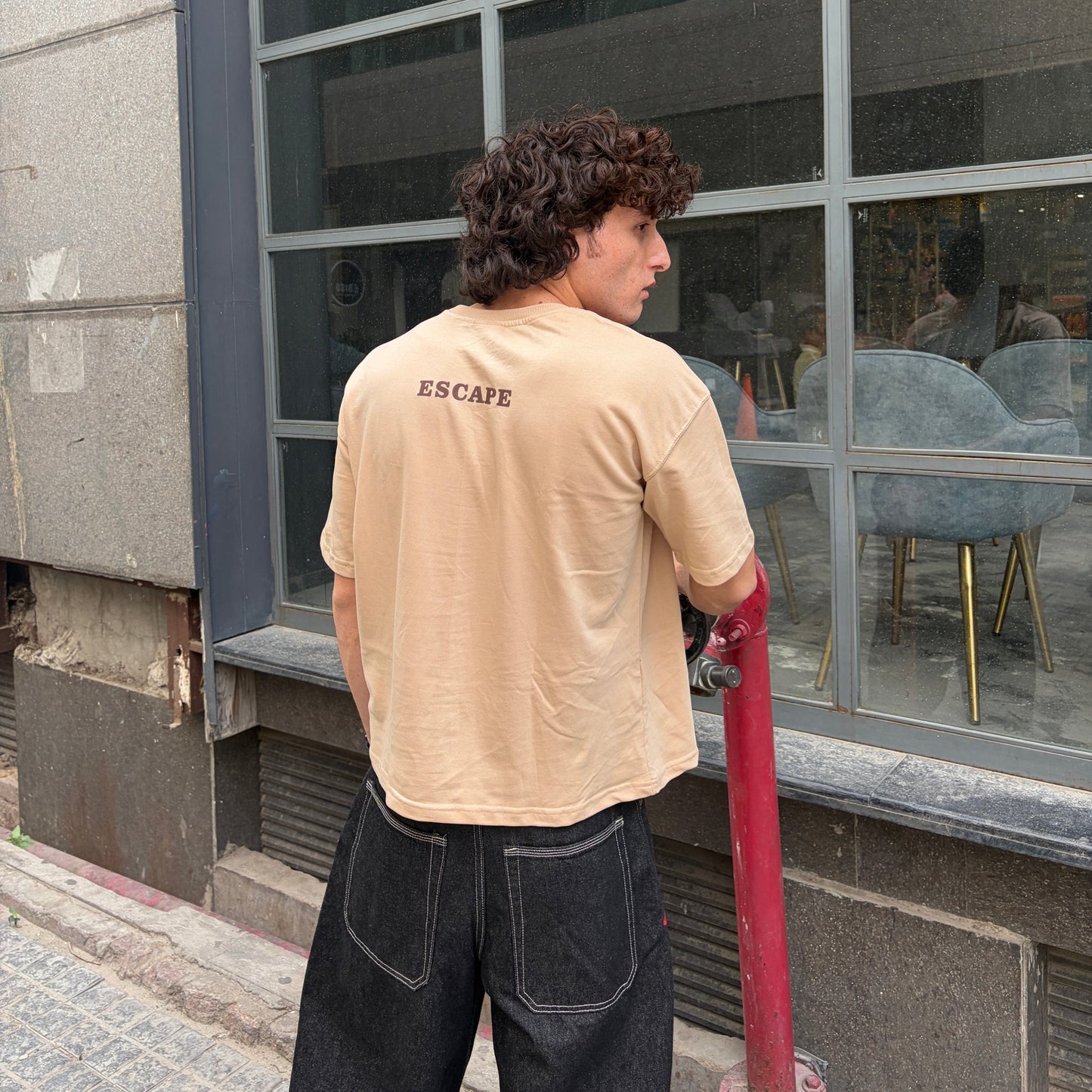 Person wearing a beige t-shirt with 'ESCAPE' printed on the back, standing outside a building with large windows.