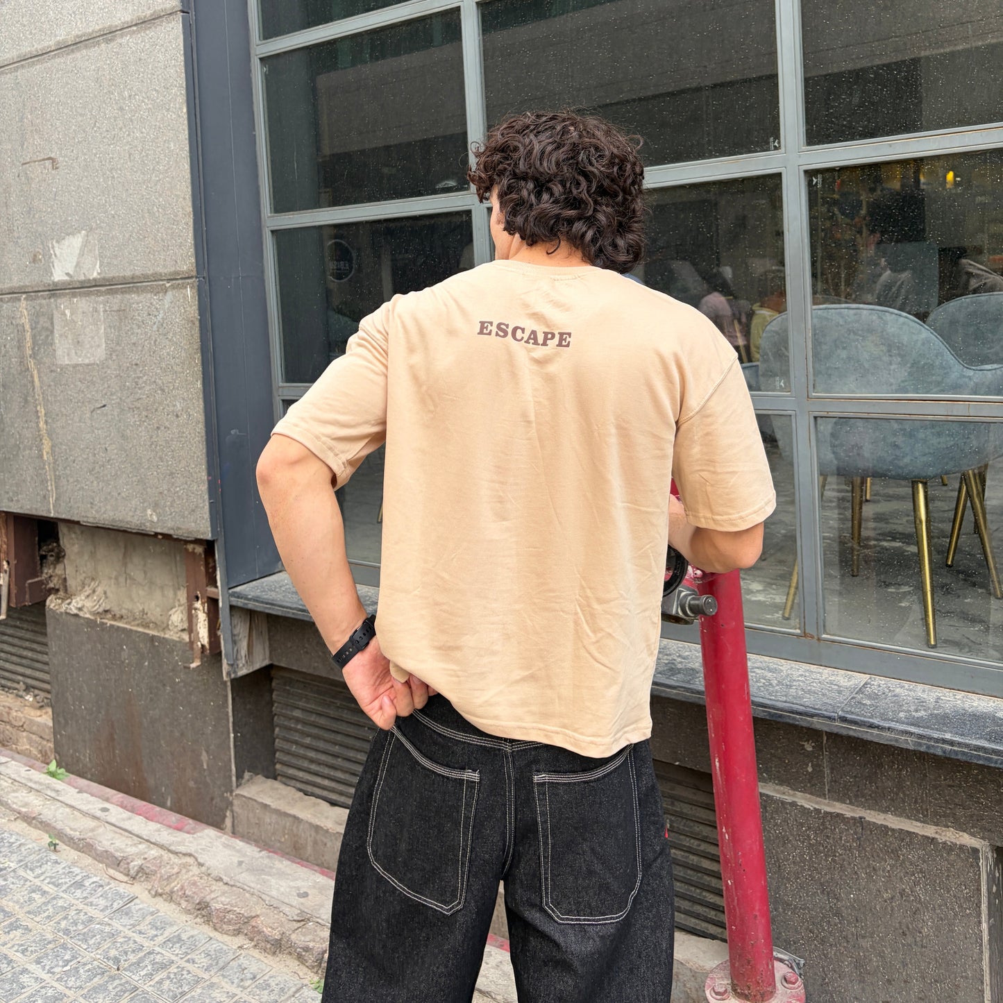 Man wearing a beige t-shirt with 'ESCAPE' printed on the back, standing outside a building.