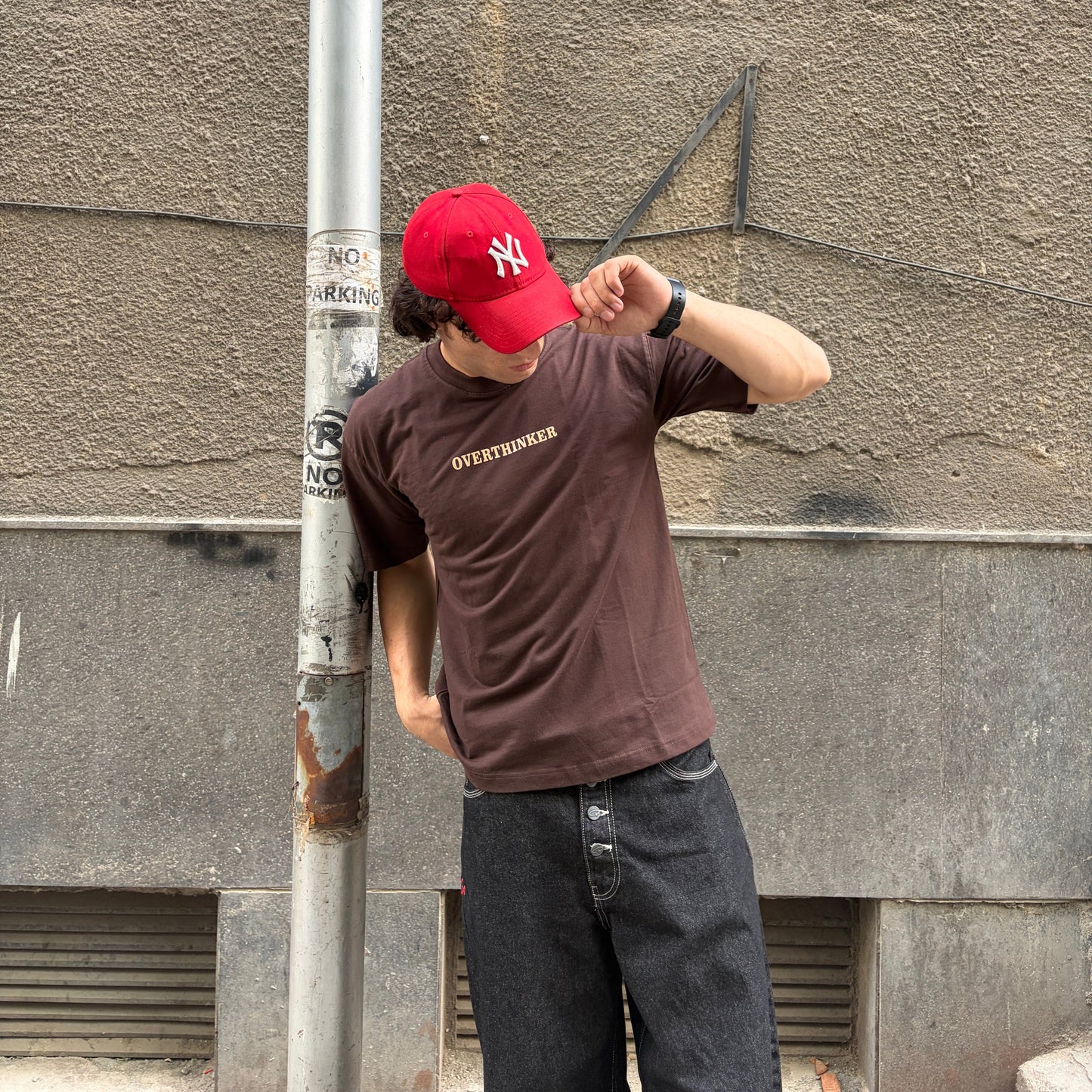 Person wearing a red cap and brown t-shirt leaning against a wall.