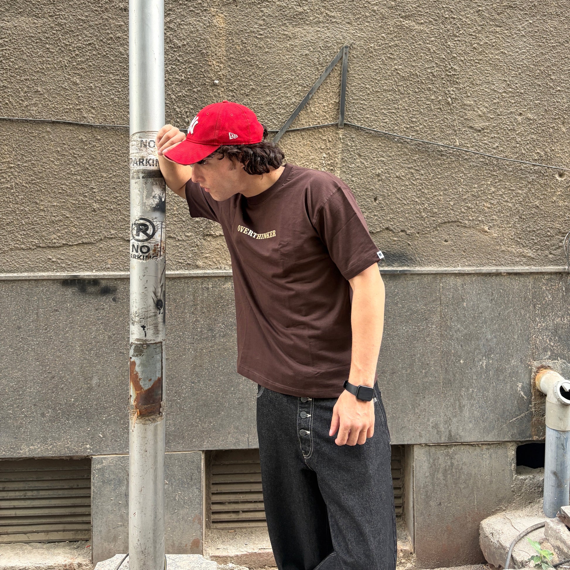 Man wearing a red cap and brown t-shirt leaning against a wall.