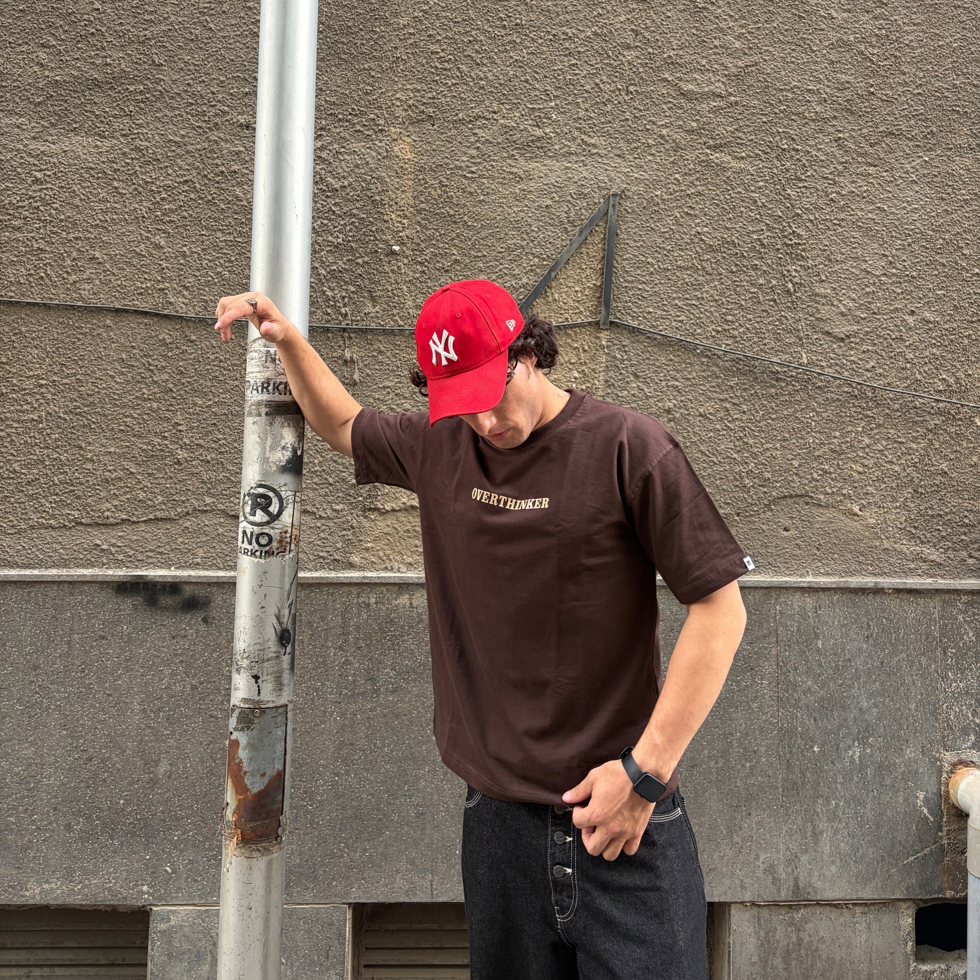Person wearing a red cap and brown t-shirt leaning against a pole with a gray wall background